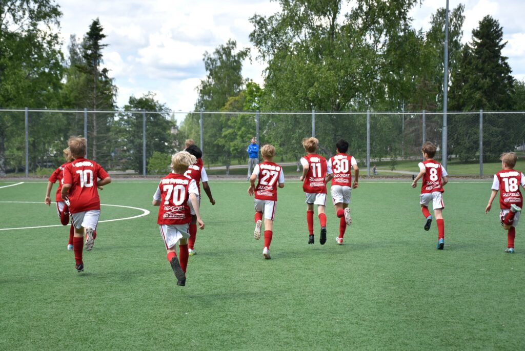 lapset juoksevat jalkapallokentällä kamerasta poispäin.