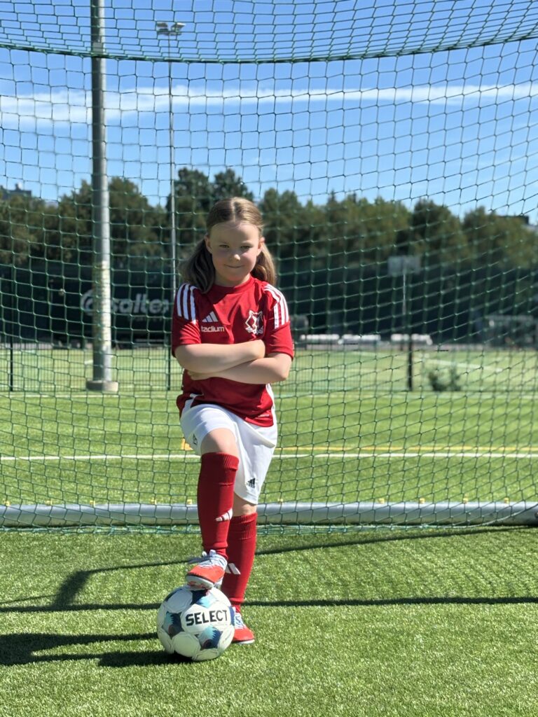 jalkapallo pelaaja tyttö maalin edessä jalka pallon päällä ja iloinen hymy kasvoilla.